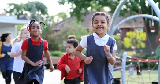 Group of primary school children smiling and running together on a playground during The Daily Mile, with climbing equipment and trees in the background.