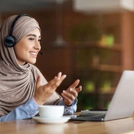 Smiling woman in a hijab wearing a headset, gesturing during a video call on her laptop in a warm, modern setting.