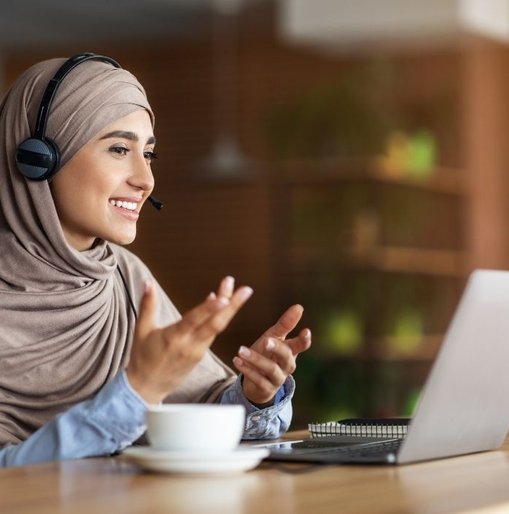 Smiling woman in a hijab wearing a headset, gesturing during a video call on her laptop in a warm, modern setting.
