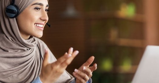 Smiling woman in a hijab wearing a headset, gesturing during a video call on her laptop in a warm, modern setting.