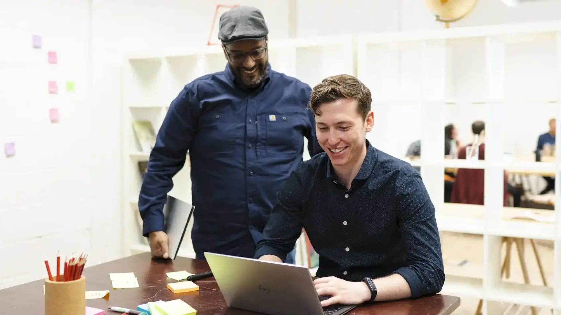 Senior backend software engineers Wayne and Will working together in a bright, creative office space—Wayne standing and smiling as Will types on a laptop, surrounded by colourful sticky notes and pens on the desk.
