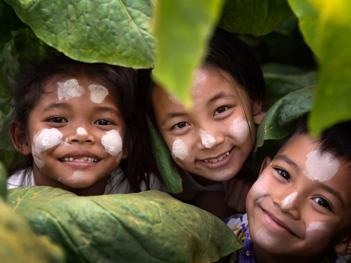 Three children with painted faces smile joyfully while peeking through large green leaves.