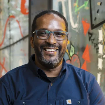 Wayne smiling in a navy shirt, seated in front of a brightly painted wall in a modern, creative indoor space.