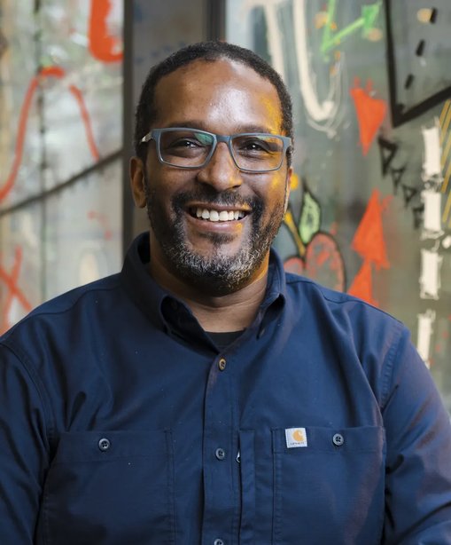 Wayne smiling in a navy shirt, seated in front of a brightly painted wall in a modern, creative indoor space.