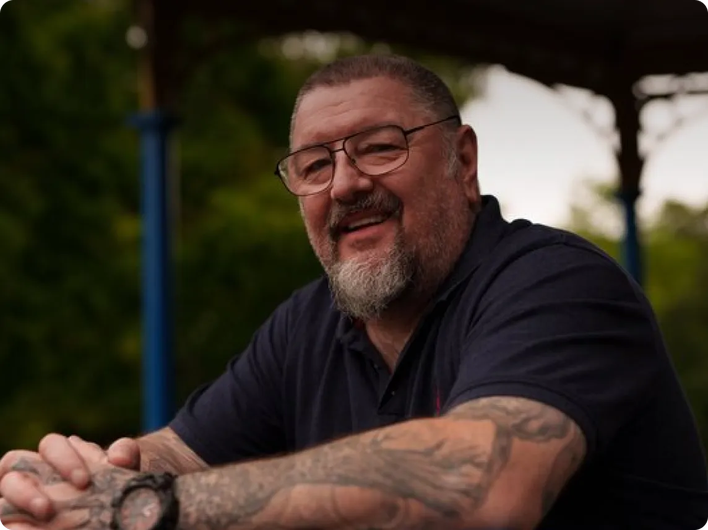 Smiling older man with glasses and tattoos on both arms, sitting outdoors under a shaded structure with greenery in the background.