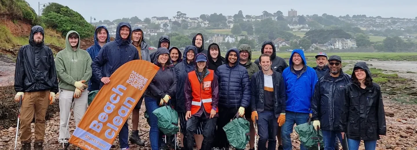 Group of Giant Digital team members smiling and posing together in rain gear during a beach clean event, holding litter pickers and an orange Marine Conservation Society banner.