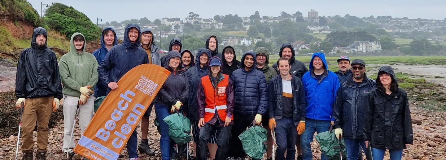 Group of Giant Digital team members smiling and posing together in rain gear during a beach clean event, holding litter pickers and an orange Marine Conservation Society banner.