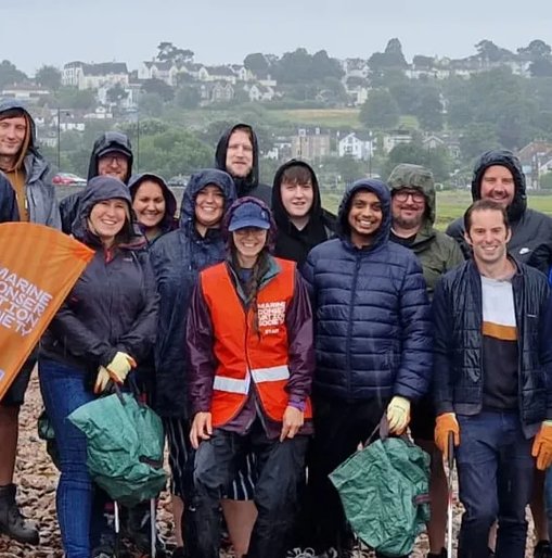 Group of Giant Digital team members smiling and posing together in rain gear during a beach clean event, holding litter pickers and an orange Marine Conservation Society banner.