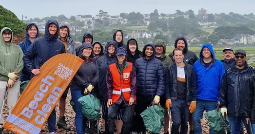 Group of Giant Digital team members smiling and posing together in rain gear during a beach clean event, holding litter pickers and an orange Marine Conservation Society banner.