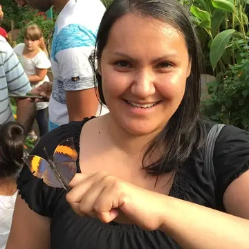 Amanda smiling and holding out her hand as a butterfly perches on her finger, surrounded by greenery in a busy butterfly house.