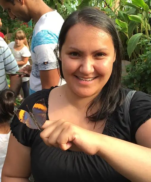 Amanda smiling and holding out her hand as a butterfly perches on her finger, surrounded by greenery in a busy butterfly house.