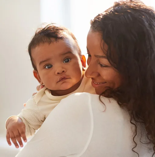A mother smiles lovingly at her baby while holding them close in a softly lit home setting. The baby looks calmly toward the camera.