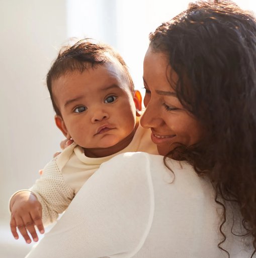 A mother smiles lovingly at her baby while holding them close in a softly lit home setting. The baby looks calmly toward the camera.