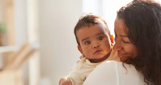 A mother smiles lovingly at her baby while holding them close in a softly lit home setting. The baby looks calmly toward the camera.