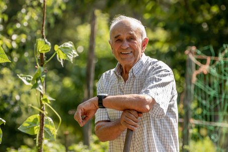 An elderly man smiling and leaning on a gardening tool in a lush, green garden on a sunny day.