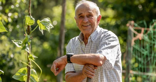 An elderly man smiling and leaning on a gardening tool in a lush, green garden on a sunny day.