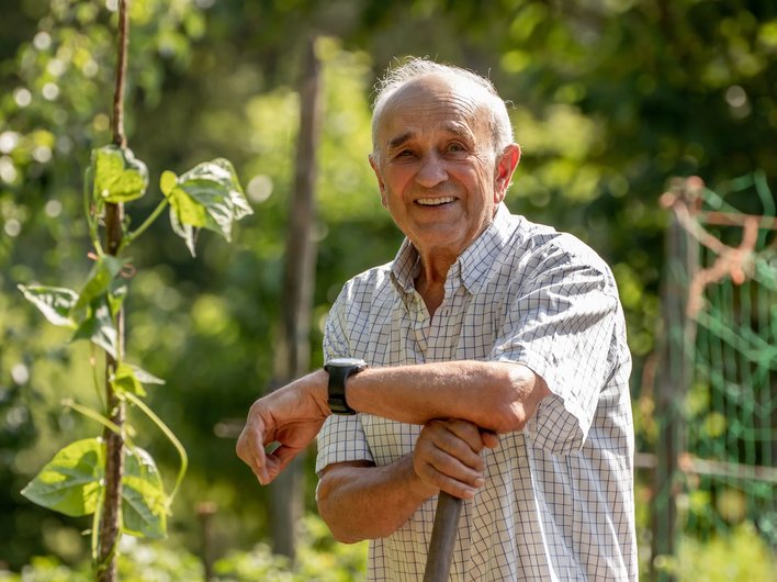 An elderly man smiling and leaning on a gardening tool in a lush, green garden on a sunny day.