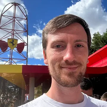 Tom taking a selfie outside, smiling beneath a blue sky with scattered clouds.