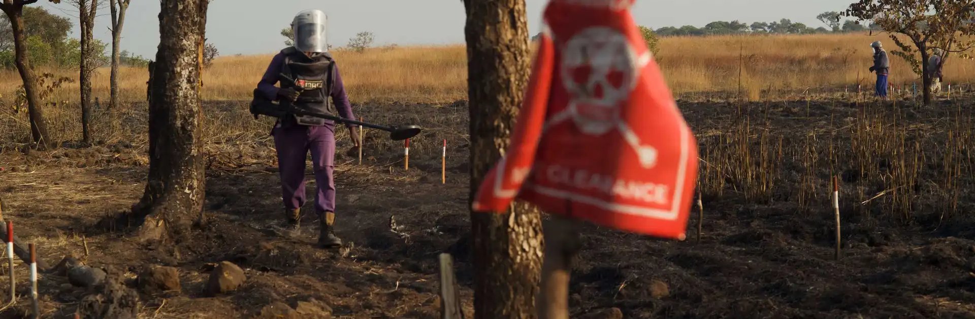 A deminer wearing protective gear and carrying detection equipment walks through a marked minefield, with a warning sign in the foreground and others working in the background.