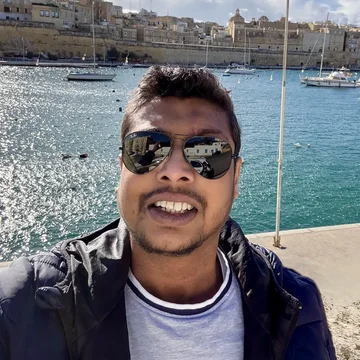 Vimal smiles in a selfie by a sparkling harbour, wearing sunglasses and a navy jacket, with a historic cityscape and moored boats in the background.
