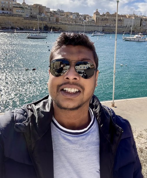 Vimal smiles in a selfie by a sparkling harbour, wearing sunglasses and a navy jacket, with a historic cityscape and moored boats in the background.
