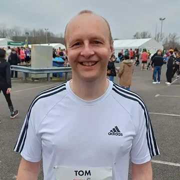 Tom smiling after a run, wearing a race number and white running shirt at a large outdoor running event.