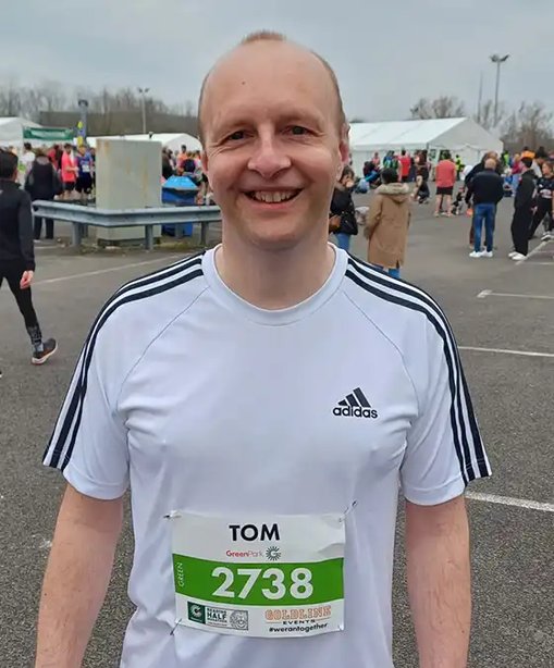 Tom smiling after a run, wearing a race number and white running shirt at a large outdoor running event.