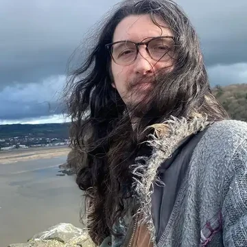 Cael taking a selfie with long hair blowing in the wind, standing on a cliffside overlooking a beach and cloudy sky.