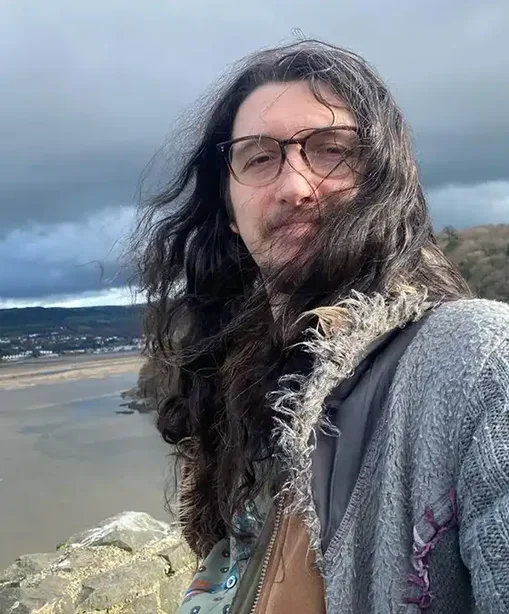 Cael taking a selfie with long hair blowing in the wind, standing on a cliffside overlooking a beach and cloudy sky.