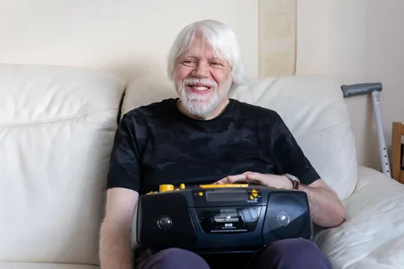 Older man with white hair and beard smiling while sitting on a cream sofa, holding an accessible DAB radio, with a walking aid visible in the background.