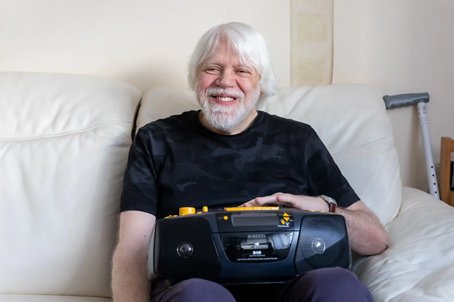 Older man with white hair and beard smiling while sitting on a cream sofa, holding an accessible DAB radio, with a walking aid visible in the background.