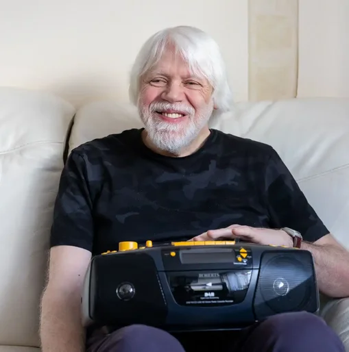 Older man with white hair and beard smiling while sitting on a cream sofa, holding an accessible DAB radio, with a walking aid visible in the background.