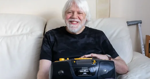 Older man with white hair and beard smiling while sitting on a cream sofa, holding an accessible DAB radio, with a walking aid visible in the background.