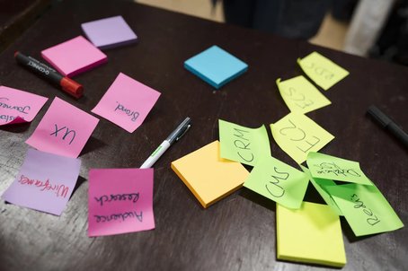 Assorted colourful sticky notes scattered across a dark table, featuring handwritten words like 'UX', 'CRM', 'CMS', and 'Audience Research', alongside pens and highlighters.