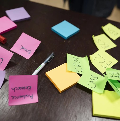 Assorted colourful sticky notes scattered across a dark table, featuring handwritten words like 'UX', 'CRM', 'CMS', and 'Audience Research', alongside pens and highlighters.