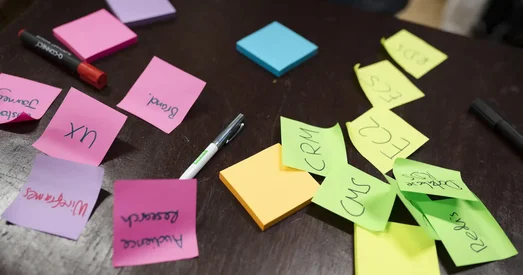 Assorted colourful sticky notes scattered across a dark table, featuring handwritten words like 'UX', 'CRM', 'CMS', and 'Audience Research', alongside pens and highlighters.