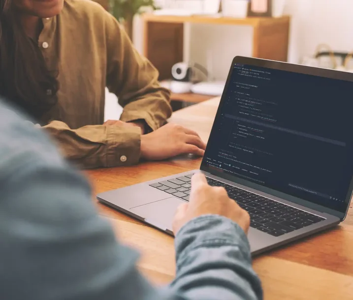 Two people sitting at a table reviewing code on a laptop screen, with one person pointing at lines of Python in a development environment.