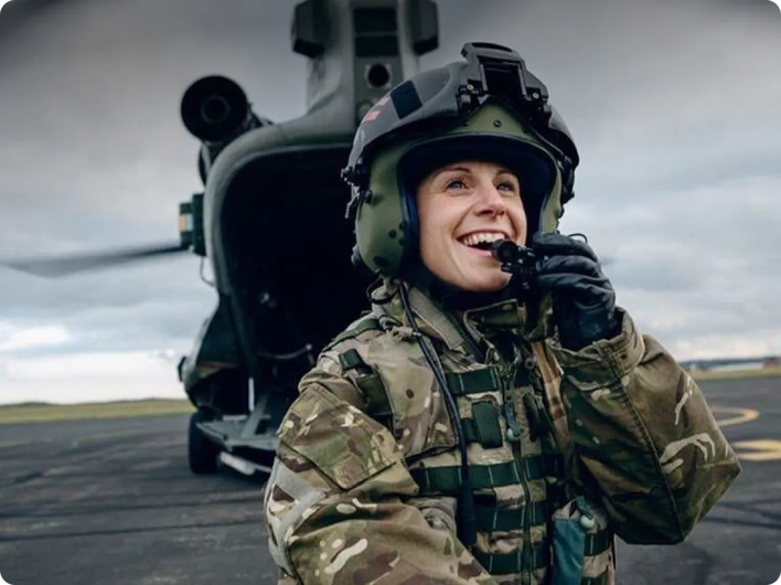 Smiling female soldier in camouflage uniform and helmet speaking into a radio, standing in front of a military helicopter on a tarmac.