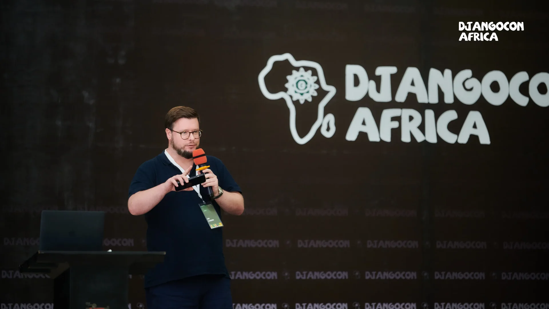 Jon speaking on stage at DjangoCon Africa, holding a microphone in front of a backdrop featuring the event’s logo and an outline of the African continent.
