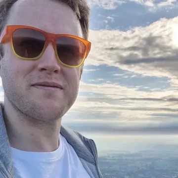 Jake stands on the Malvern Hills wearing orange sunglasses and a white T-shirt that reads “good good good good double good”, with a scenic view stretching out behind him under a dramatic cloudy sky.