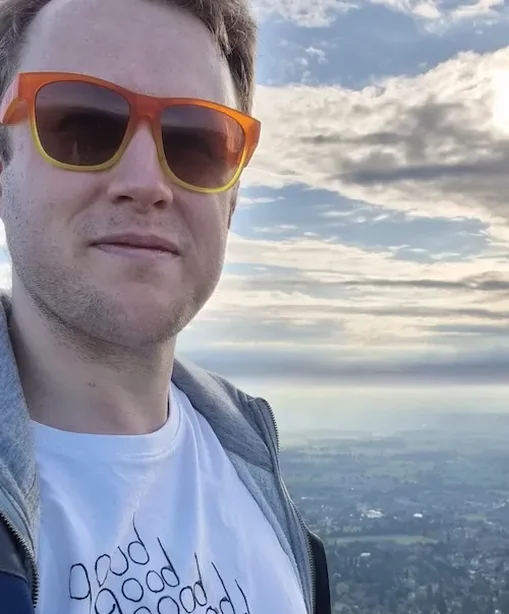 Jake stands on the Malvern Hills wearing orange sunglasses and a white T-shirt that reads “good good good good double good”, with a scenic view stretching out behind him under a dramatic cloudy sky.