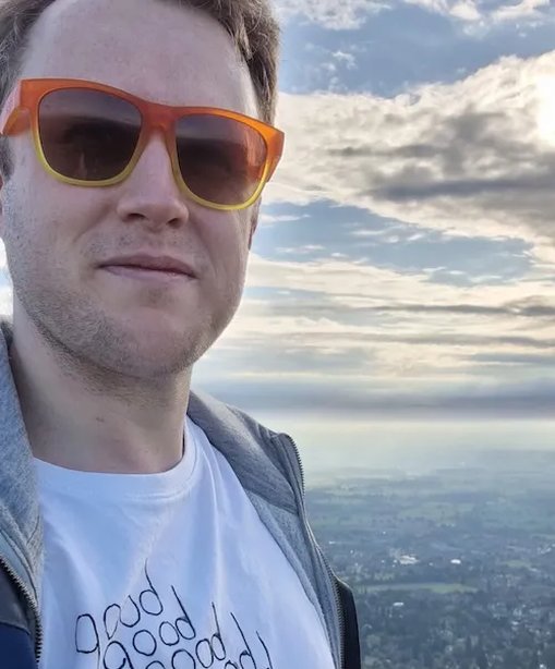 Jake stands on the Malvern Hills wearing orange sunglasses and a white T-shirt that reads “good good good good double good”, with a scenic view stretching out behind him under a dramatic cloudy sky.