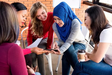 A group of women sitting in a circle, smiling and collaborating over a tablet during an informal meeting.