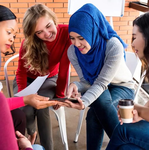 A group of women sitting in a circle, smiling and collaborating over a tablet during an informal meeting.