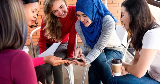 A group of women sitting in a circle, smiling and collaborating over a tablet during an informal meeting.