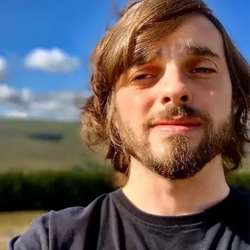 Karl taking a selfie under a bright blue sky, wearing a black t-shirt with hills and scattered clouds in the background.