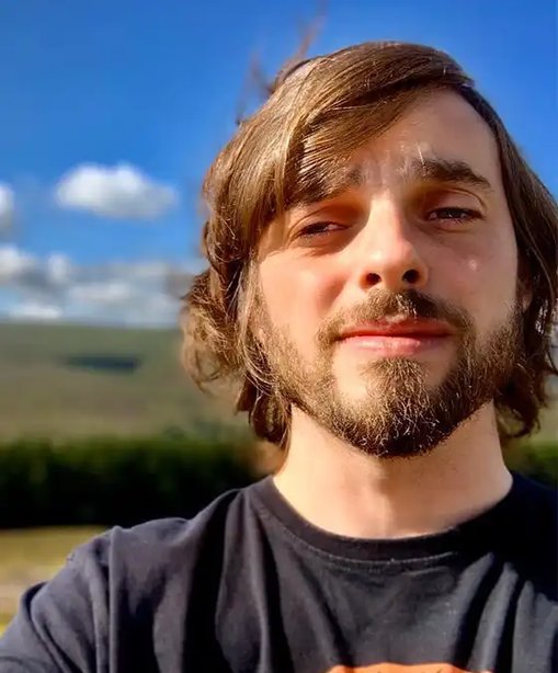 Karl taking a selfie under a bright blue sky, wearing a black t-shirt with hills and scattered clouds in the background.