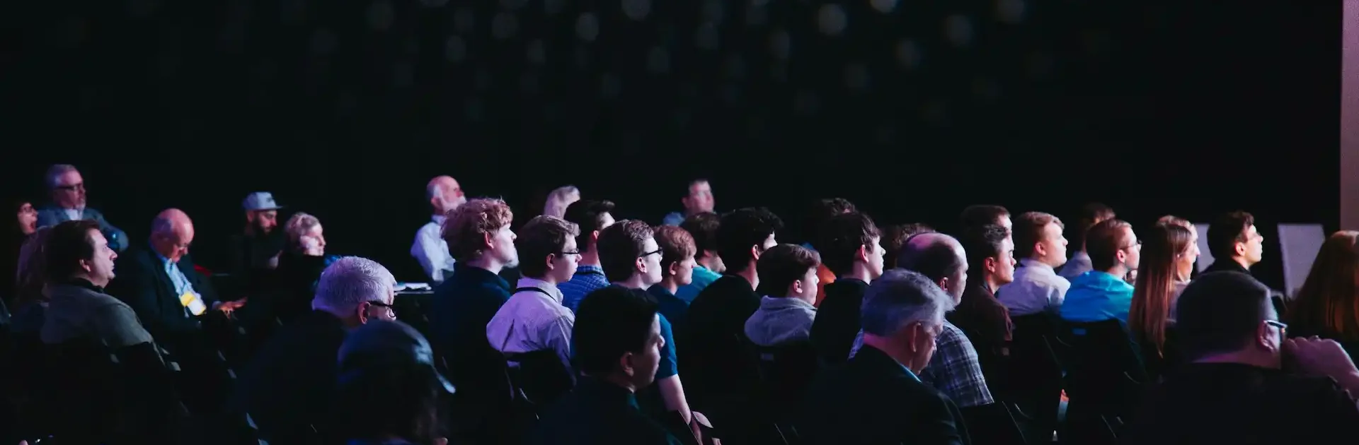 Audience seated in a dark auditorium, attentively watching a presentation or speaker under spotlights creating a patterned light effect on the wall.