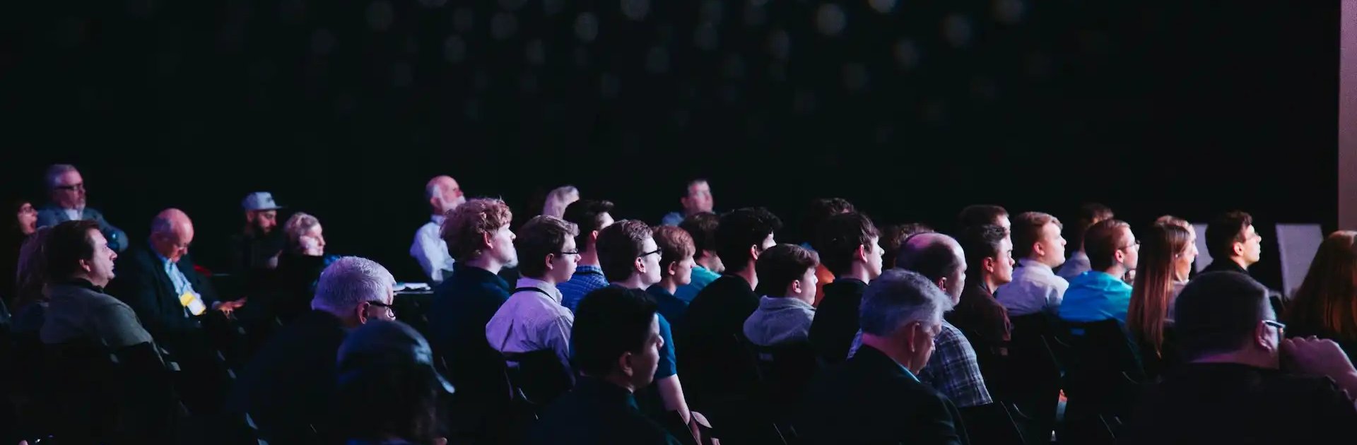 Audience seated in a dark auditorium, attentively watching a presentation or speaker under spotlights creating a patterned light effect on the wall.