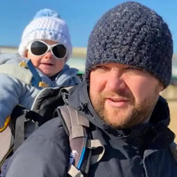 Alex bundled up in winter clothes with his baby on his back, both wearing woolly hats and sunglasses on a sunny day out.